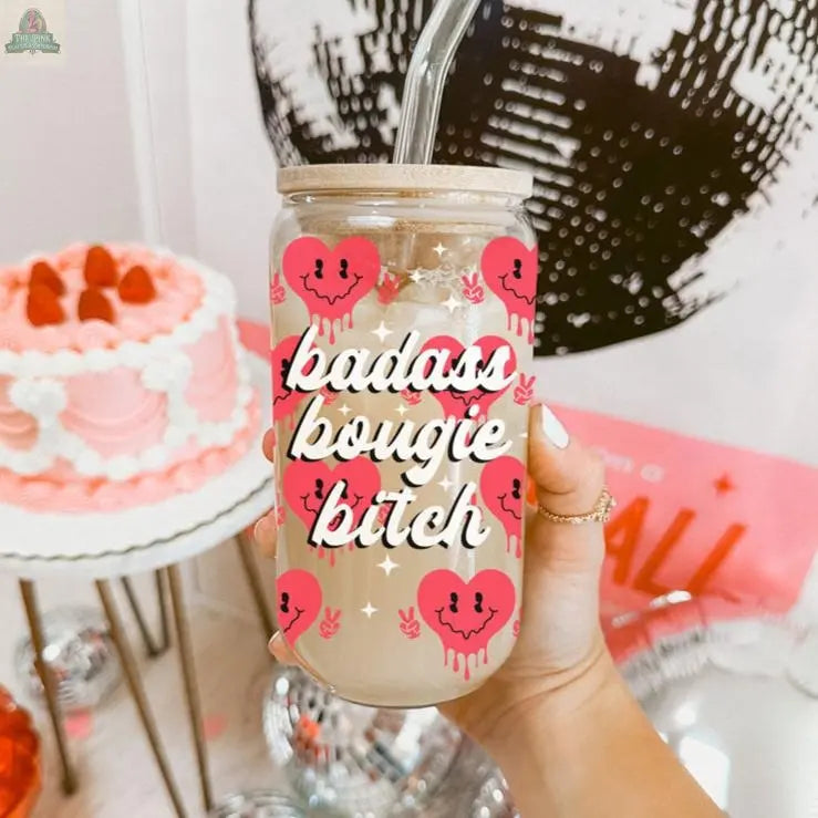 A hand holds a Badass Bougie 20oz glass cup with bamboo lid and straw, decorated with pink hearts and bold lettering. In the background are a pink and white cake and abstract black and white wall art.