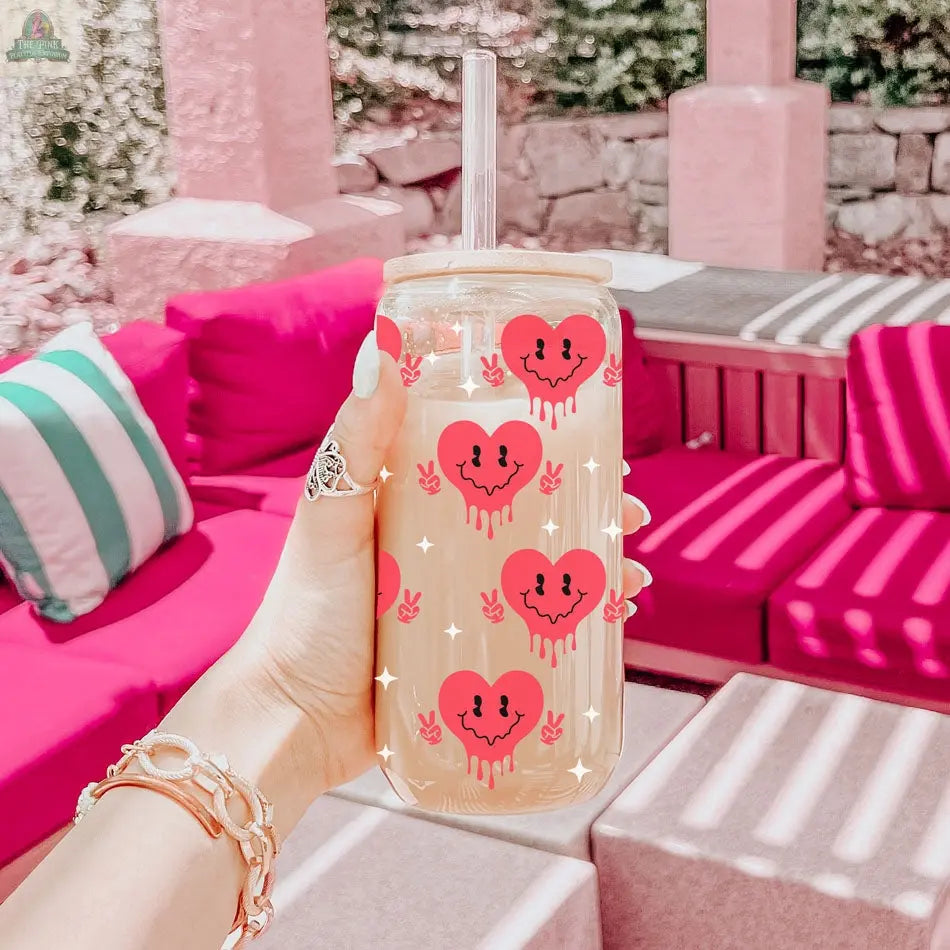 A hand holds the Badass Bougie 20oz glass cup filled with iced coffee, featuring cute red heart faces; pink outdoor seating and striped pillows are visible in the background.