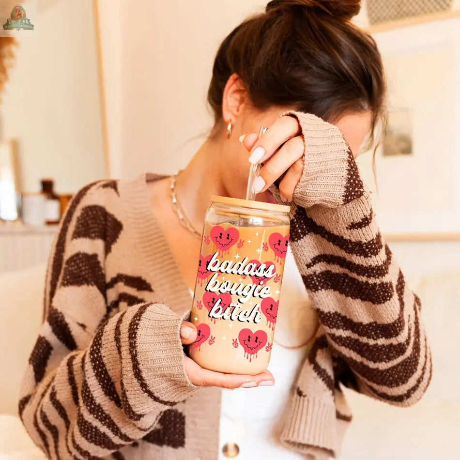 A woman in a striped cardigan relaxes in a cozy, softly lit room, holding a Badass Bougie 20oz glass cup with a straw and partially covering her face with her hand.