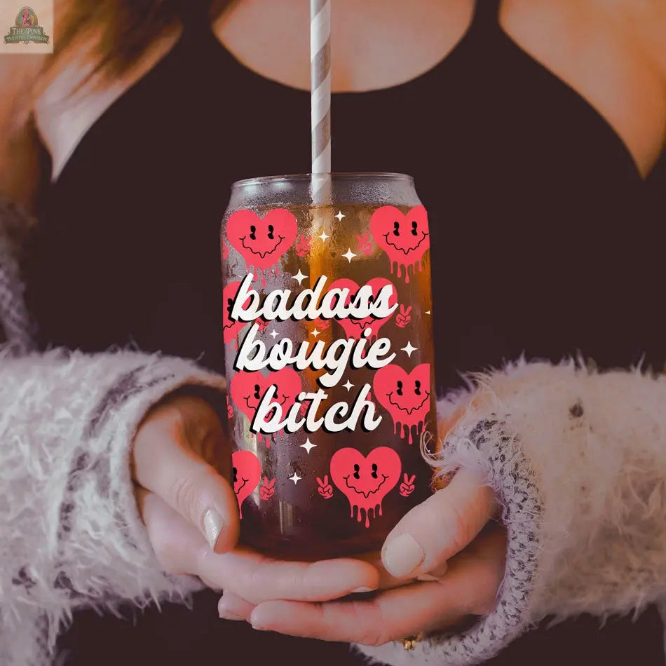 A person in a black top and fuzzy sweater holds a 20oz Badass Bougie cup decorated with pink smiley hearts and bold white and pink lettering, featuring a striped straw for stylish sipping.