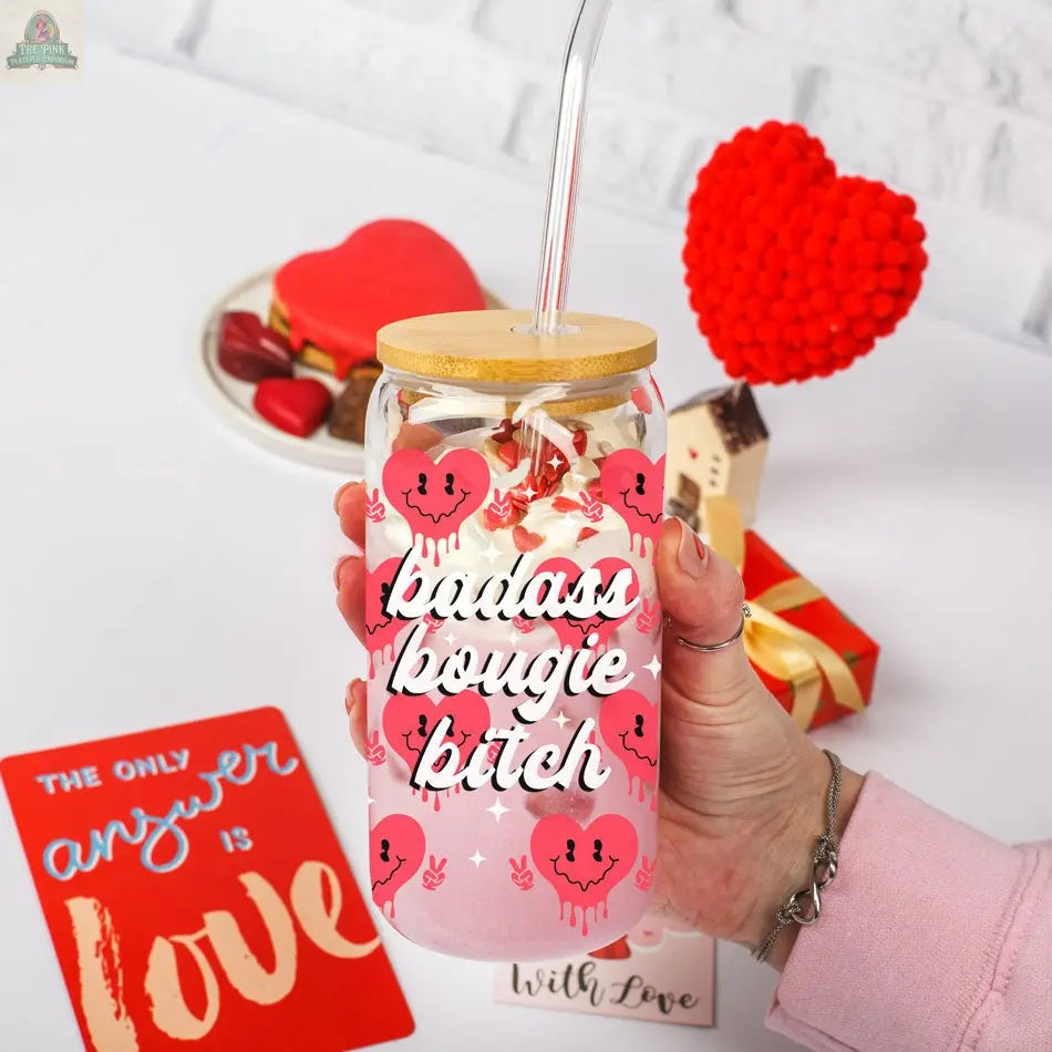 A hand holds the Badass Bougie 20oz glass cup with a bamboo lid and straw, featuring pink hearts. In the background are heart-shaped treats, a gift box, and a red card that says, "The only answer is love.