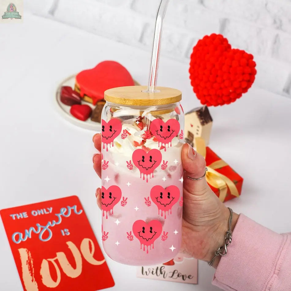 A hand holds a Badass Bougie 20oz glass cup with cute heart faces, filled with pink drink and whipped cream. In the background are heart-shaped treats, a gift box, and a red card reading "The only answer is love.