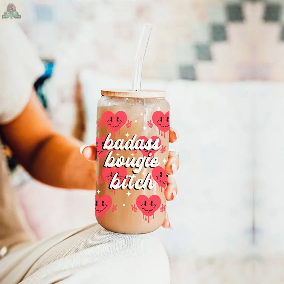A person holds a 20oz Badass Bougie glass cup with a straw, featuring pink cartoon hearts and bold white script, set against a softly blurred background.