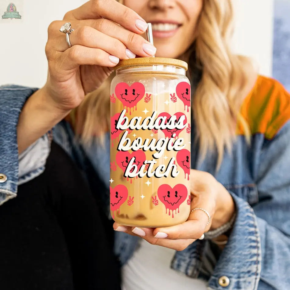 A person holds a Badass Bougie 20oz glass cup with a bamboo lid and straw, featuring pink broken hearts and bold white text. The vibrant cup stands out against their denim jacket and light hair.
