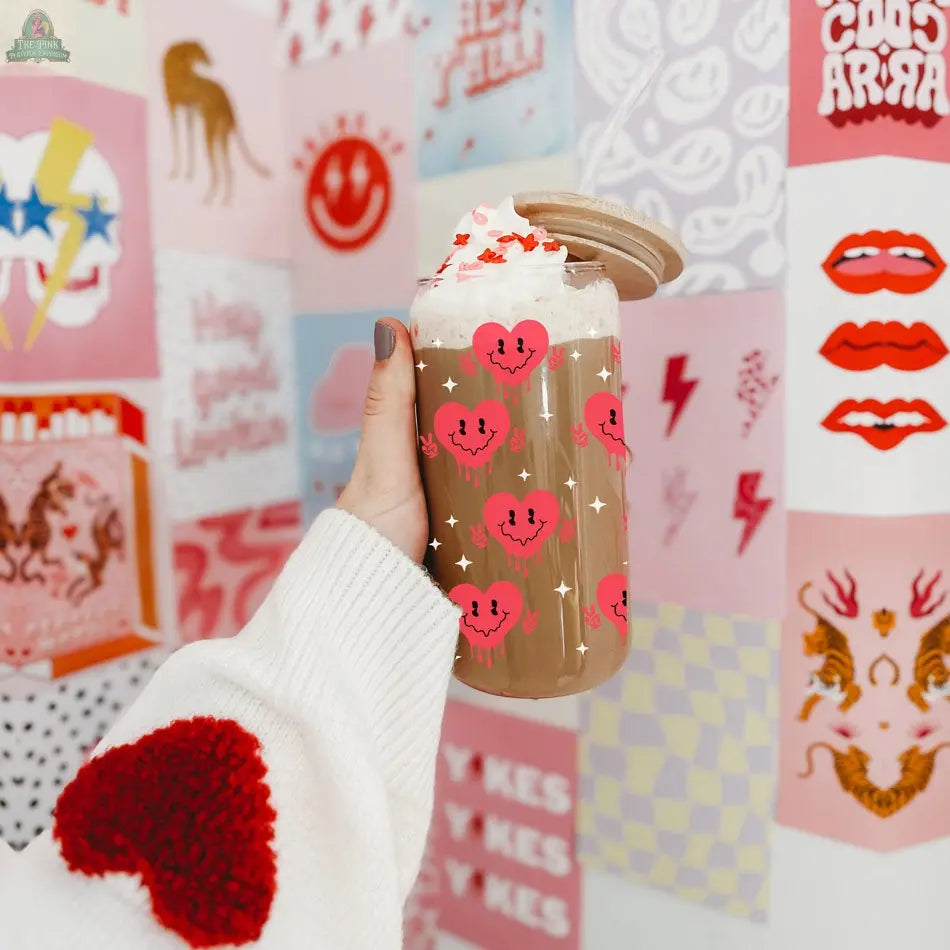 A hand in a white sweater with a red heart holds the Badass Bougie 20oz glass, filled with iced coffee and whipped cream. The smiling red heart design pops against vibrant, graphic wall art in the background.