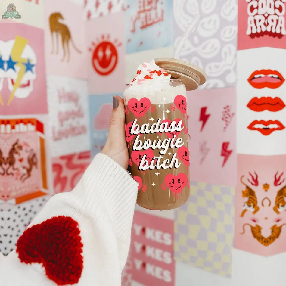A hand in a white sweater with a red heart holds a Badass Bougie 20oz glass cup filled with iced coffee and whipped cream. Pink smiley faces and bold, graphic art prints make a vibrant background for this stylish cup.