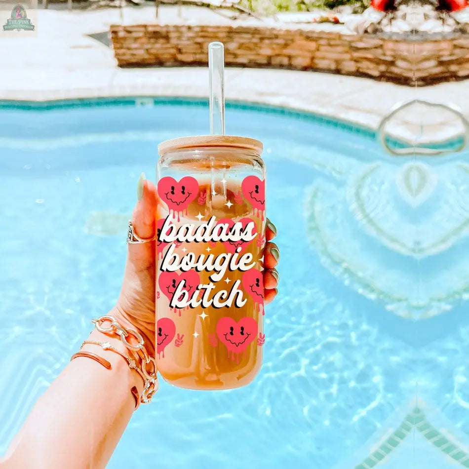 A hand with bracelets holds a 20oz Badass Bougie glass cup, decorated with pink hearts and bold white script, filled with iced coffee and a straw by the pool for a chic summer vibe.