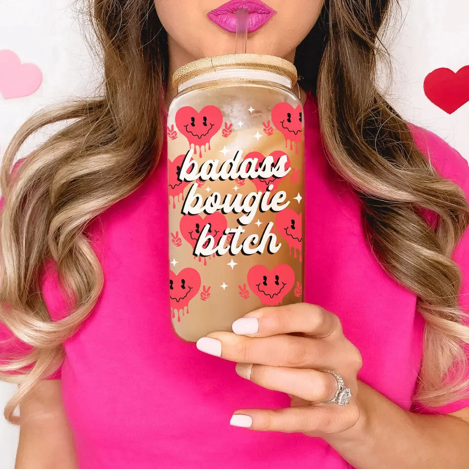 A woman in a pink shirt holds a 20oz Badass Bougie glass cup with a bamboo lid and straw, featuring red hearts and white lettering. The stylish cup complements her look as she displays a ring on her left hand.