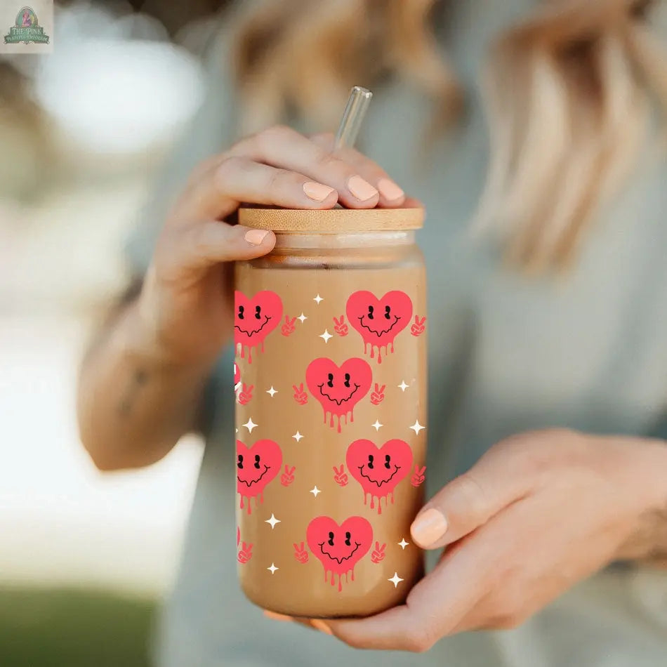 A person with light-colored nails holds a Badass Bougie 20oz bamboo lid glass cup, decorated with smiling, dripping pink hearts and white stars, filled with a light brown drink.