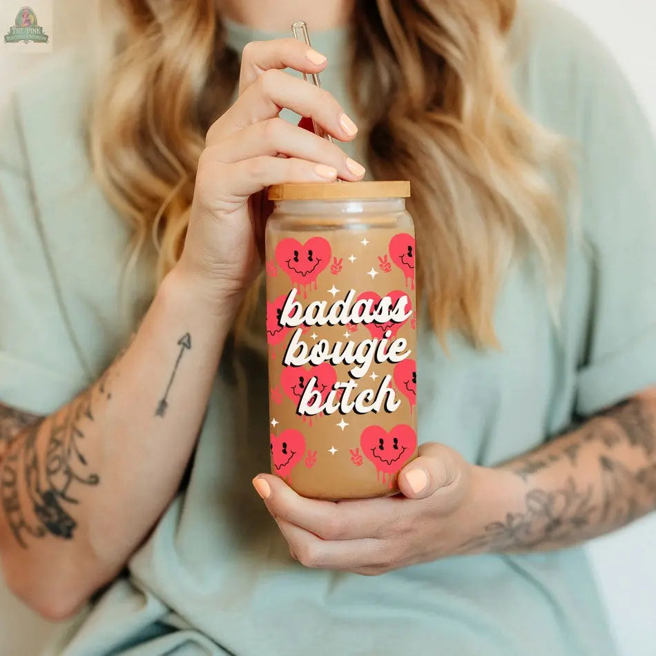 A woman with long hair and tattooed arms holds a Badass Bougie 20oz glass cup, featuring hearts, sparkles, and a colorful design, complete with a bamboo lid and straw that reflect her bold, stylish vibe.