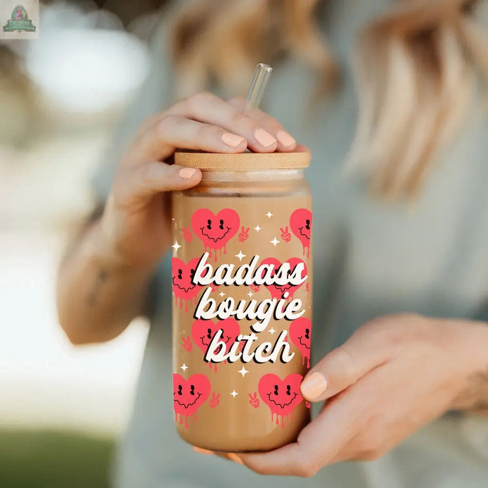 A person holds a Badass Bougie 20oz glass cup with bamboo lid and straw, decorated with pink dripping hearts and bold white script, filled with iced coffee.