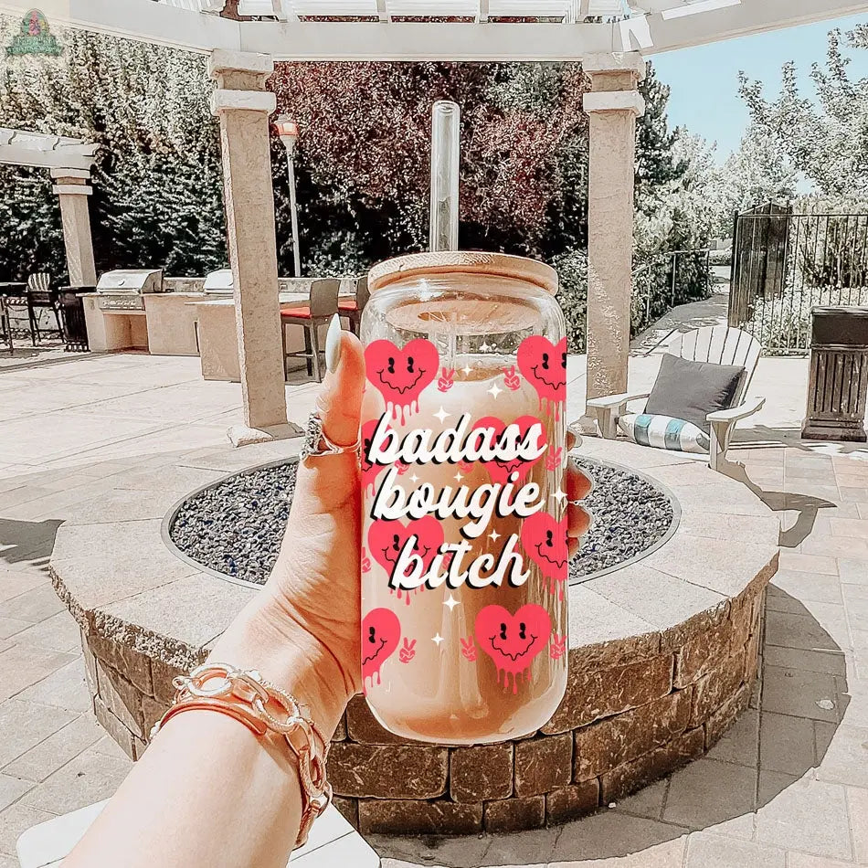 A hand with gold bracelets holds a Badass Bougie 20oz glass cup featuring pink smiley faces and a straw, set against a patio backdrop with chairs, a fire pit, and lush greenery.