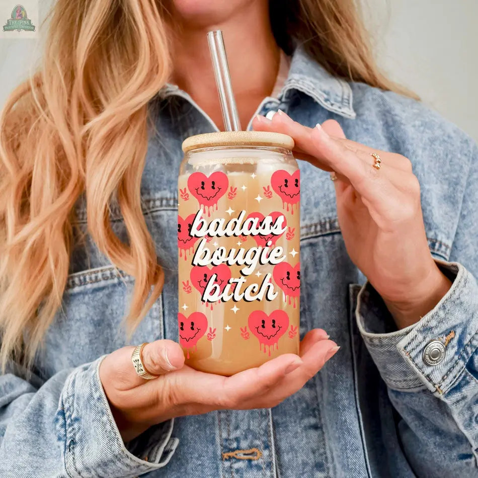 A person in a denim jacket holds a Badass Bougie 20oz glass cup with a bamboo lid and straw. The jar features pink heart designs and bold lettering, complementing their long blonde hair and stylish rings.