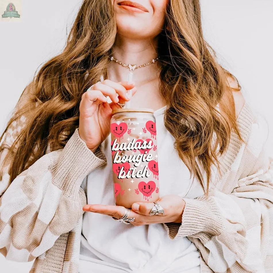 A woman with long, wavy hair holds a Badass Bougie 20oz glass cup with smiling cherries and a bamboo lid. Wearing layered necklaces and rings with a beige cardigan, she displays a fun, chic vibe.
