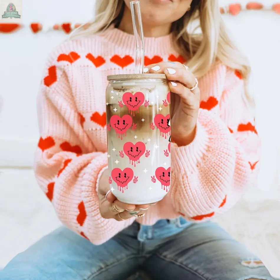 A woman in a pink sweater with red hearts sits cross-legged on the floor, enjoying iced coffee from a Badass Bougie 20oz glass cup decorated with pink smiling hearts and sipping through a clear straw.