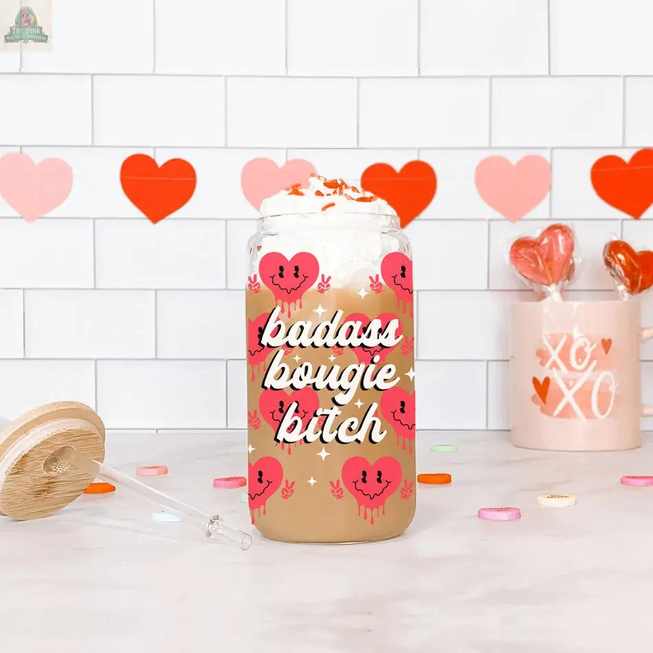 A Badass Bougie 20oz glass cup filled with iced coffee, topped with whipped cream and sprinkles, sits on a white counter. The cup features pink heart faces, while red heart decorations and candy appear in the background.