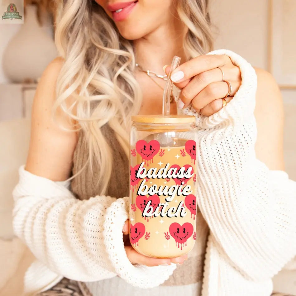 A woman with long, wavy blonde hair holds a 20oz Badass Bougie glass cup with a bamboo lid and straw, featuring cute red heart faces and bold white script. She wears a cozy white sweater.