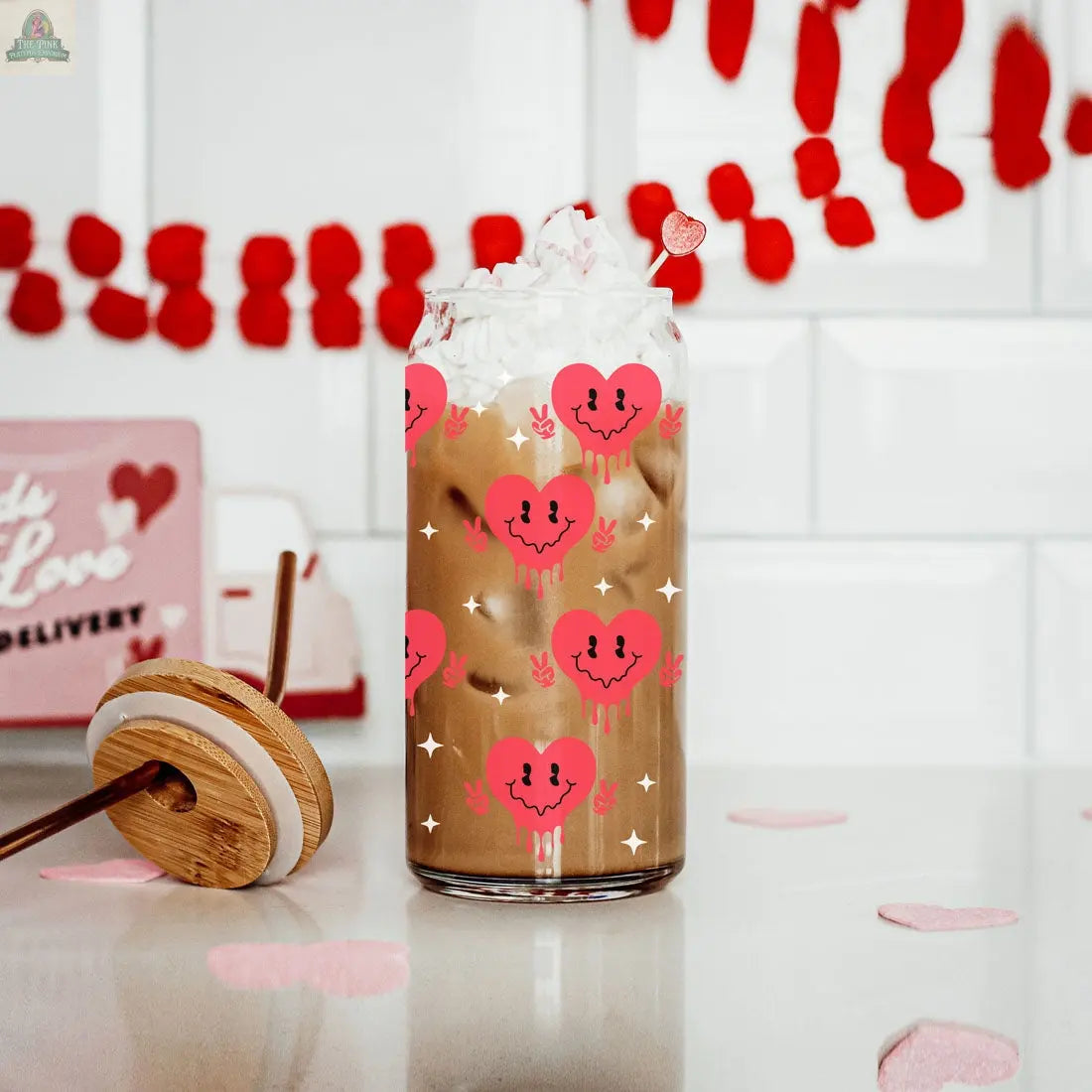 A glass of iced coffee with whipped cream served in a Badass Bougie 20oz cup, featuring pink and red smiling hearts. The background displays red garland, a pink sign, and heart-shaped felt pieces on the counter.