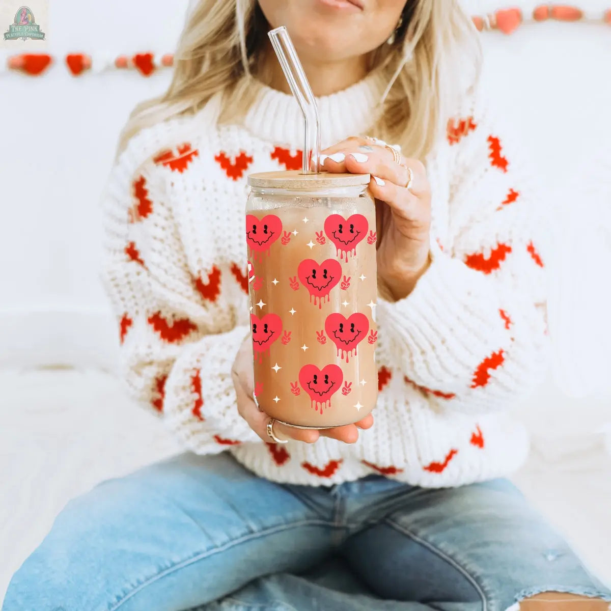A woman in a white sweater with red hearts sips iced coffee through a clear straw from the Badass Bougie 20oz glass tumbler with pink smiling heart designs and a bamboo lid, sitting cross-legged in blue jeans.