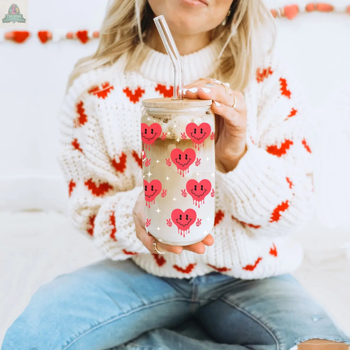 A woman in blue jeans and a white sweater with red hearts holds the Badass Bougie 20oz glass tumbler, featuring a bamboo lid, straw, and pink smiling heart designs. She sits cross-legged.