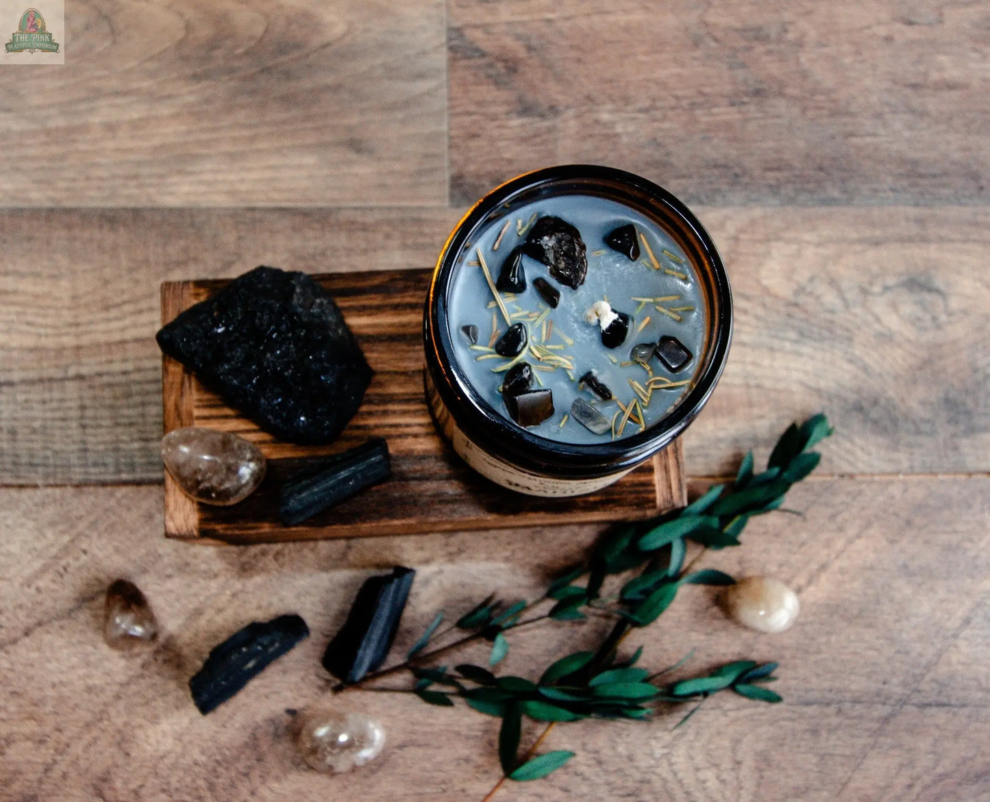 The Banish metaphysical candle in a brown glass jar with herbs and stones rests on a wooden tray, surrounded by black tourmaline, clear crystals, and green leaves atop a wooden surface.