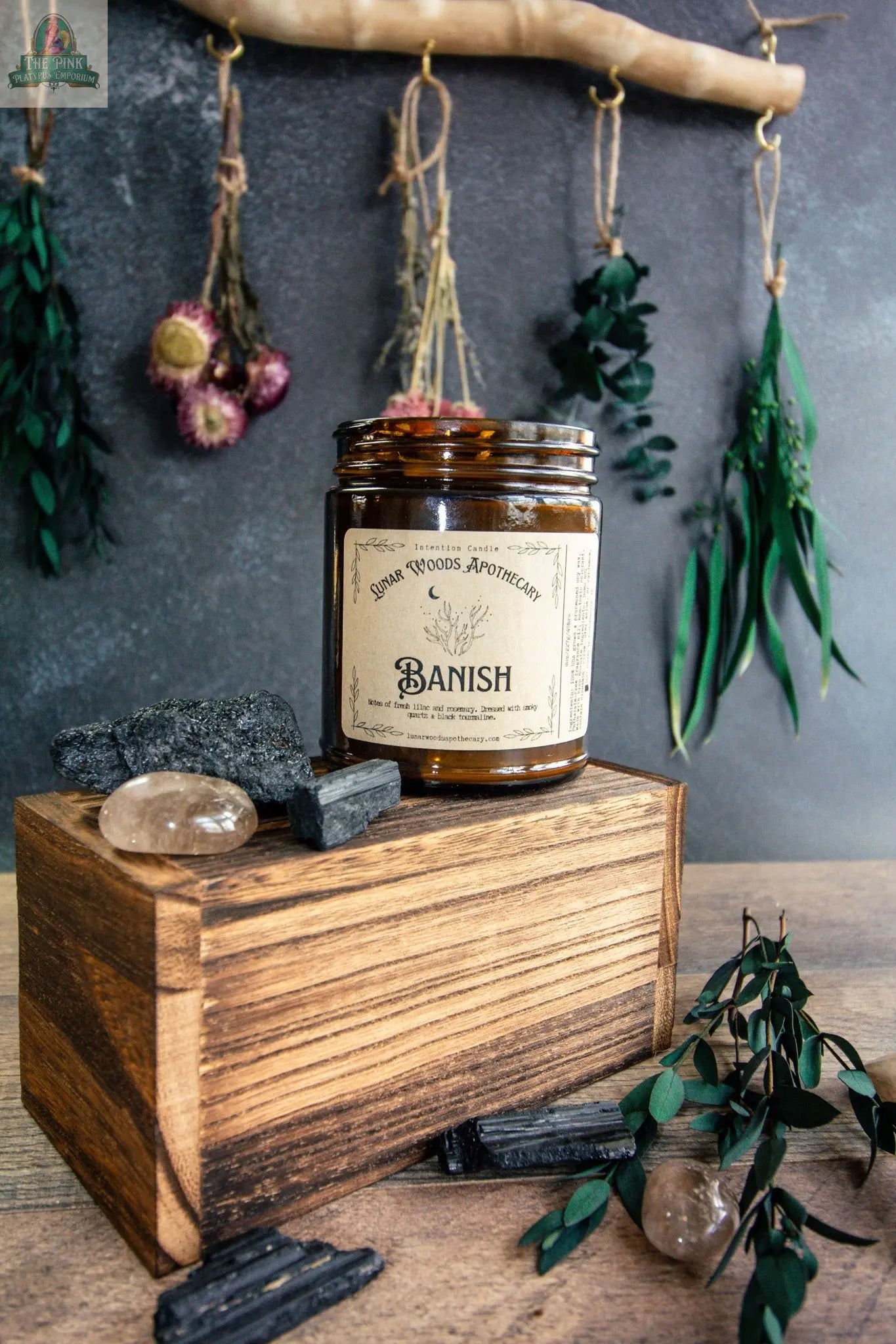 A brown glass Banish candle rests on a wooden box, surrounded by black stones, a clear crystal, and greenery; dried herbs and flowers hang in the background, creating an inviting metaphysical display.