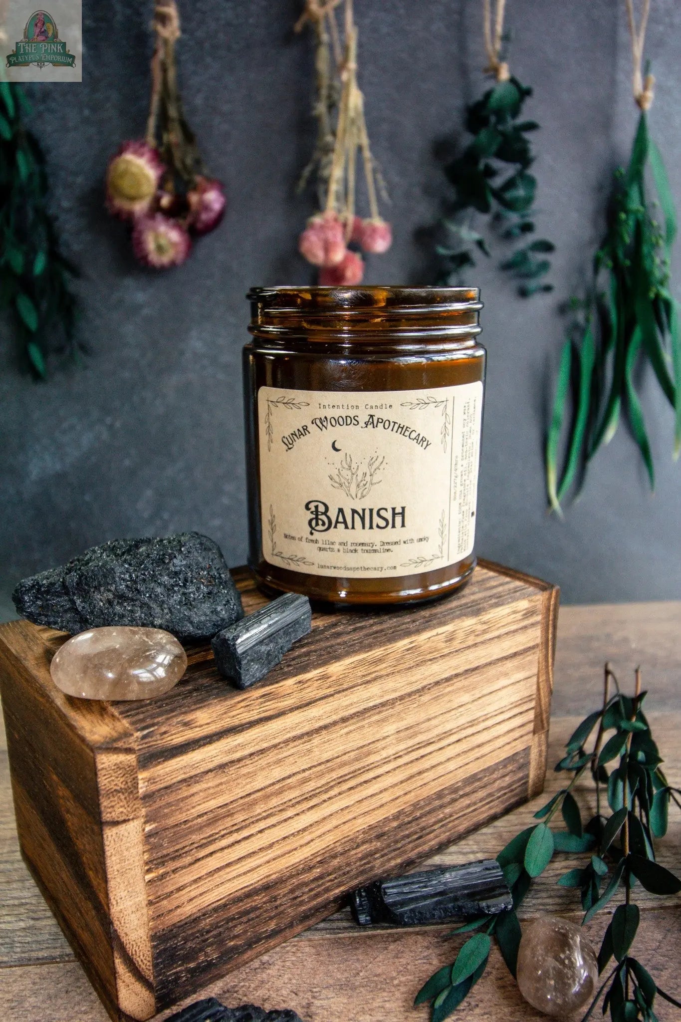 A brown glass jar labeled "Banish" rests on a wooden box amid crystals, black stones, and green leaves. Dried flowers in the background enhance the apothecary vibe—perfect for this metaphysical Banish candle to dispel negativity.