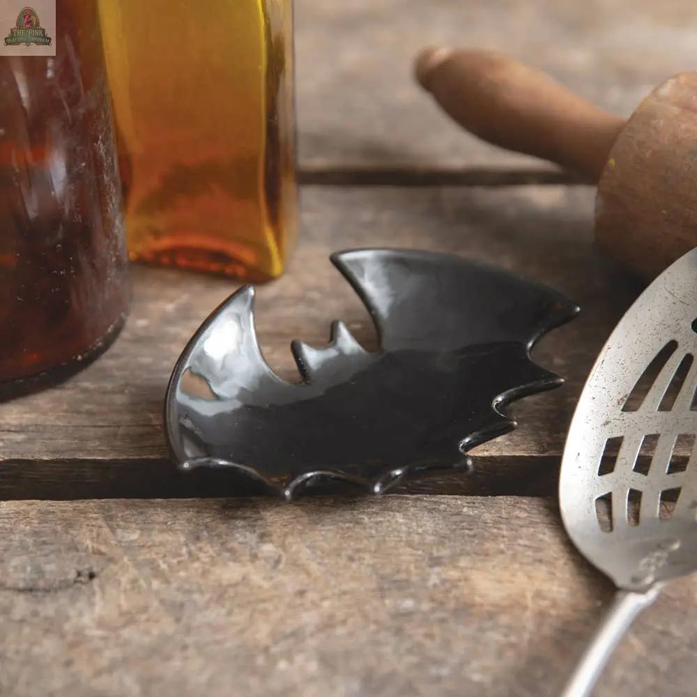 A small, glossy black Bat Spoon Rest, perfect for Halloween decor, sits on wood beside a slotted spoon, rolling pin, and amber bottles—an eye-catching addition to your porcelain kitchen essentials.