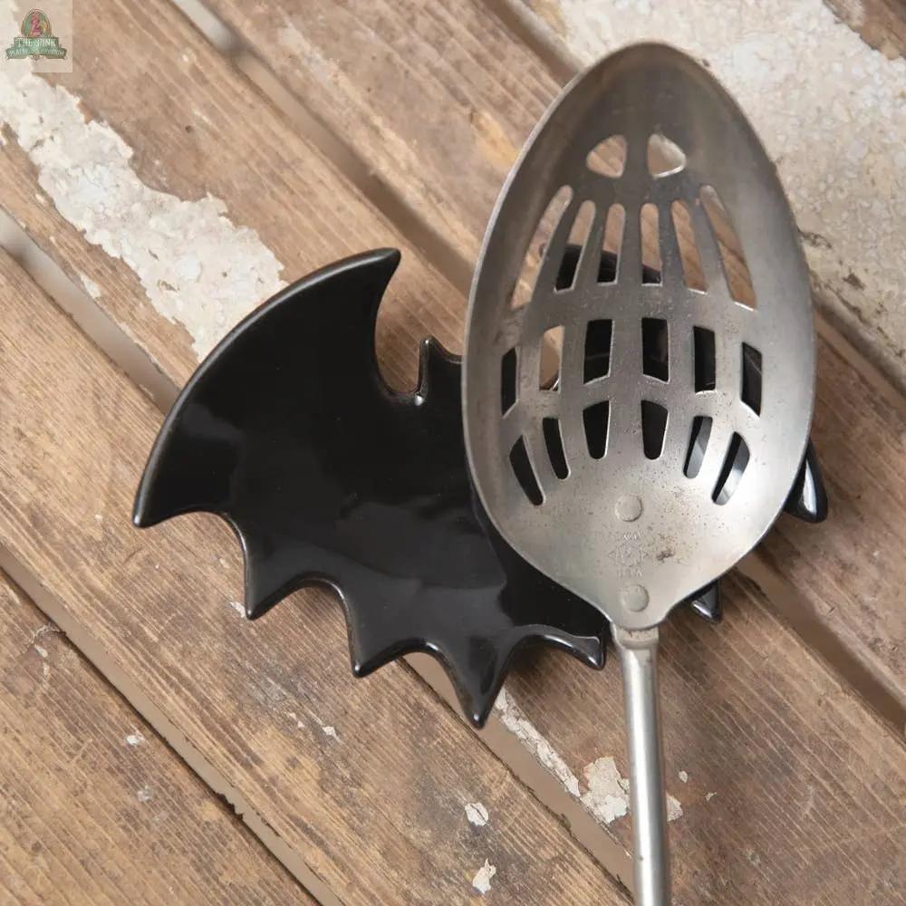 A slotted metal spoon sits on the Bat Spoon Rest, shaped like a bat, bringing whimsical Halloween flair to a rustic wooden surface with peeling paint.