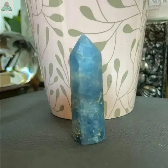 A Beautiful Natural Blue Celestite Tower from Bodhi Crystals stands upright before a light plant pot with a leafy pattern, adding calming energy to the space, with blurred greenery and objects in the background.