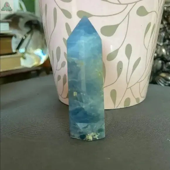 The Beautiful Natural Blue Celestite Tower from Bodhi Crystals stands upright on a gray surface before a pink floral pot, radiating calm energy—an elegant accent for any crystal collection. Blurred books and decor appear in the background.