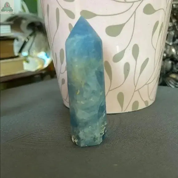 A Beautiful Natural Blue Celestite Tower from Bodhi Crystals stands upright on a gray surface before a pink vase with green leaves, bringing calming energy to your crystal collection. Books and objects appear blurred in the background.