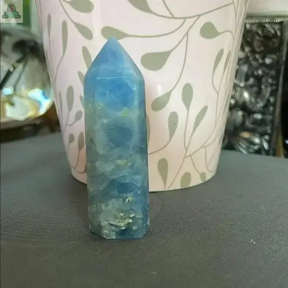A Beautiful Natural Blue Celestite Tower from Bodhi Crystals stands upright on a gray surface, in front of a white flowerpot with a light green leafy vine pattern—an ideal addition to your calming crystal collection.