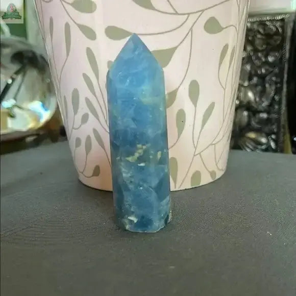 The Beautiful Natural Blue Celestite Tower by Bodhi Crystals stands upright on a gray surface before a light-colored, leafy plant pot, adding calming energy to your crystal collection with its serene blue hue. Blurred objects are visible in the background.