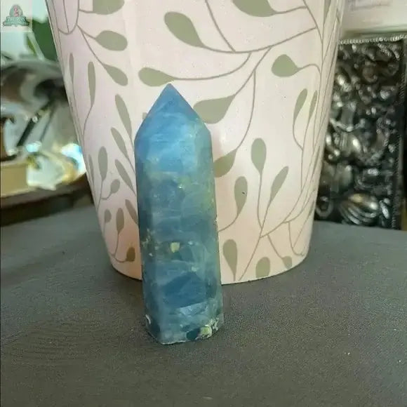 The Beautiful Natural Blue Celestite Tower from Bodhi Crystals stands upright on a gray surface before a pale pink flowerpot with green leaf patterns, radiating calming energy and making a serene addition to any collection.