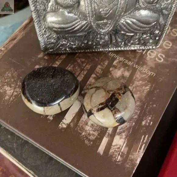 Two polished Beautiful Palm Dragon Stone Druzy stones (about 2" diameter, brown, black, and cream patterns) rest on a brown book. Behind them is a metallic object with an embossed design, possibly a seated figure.