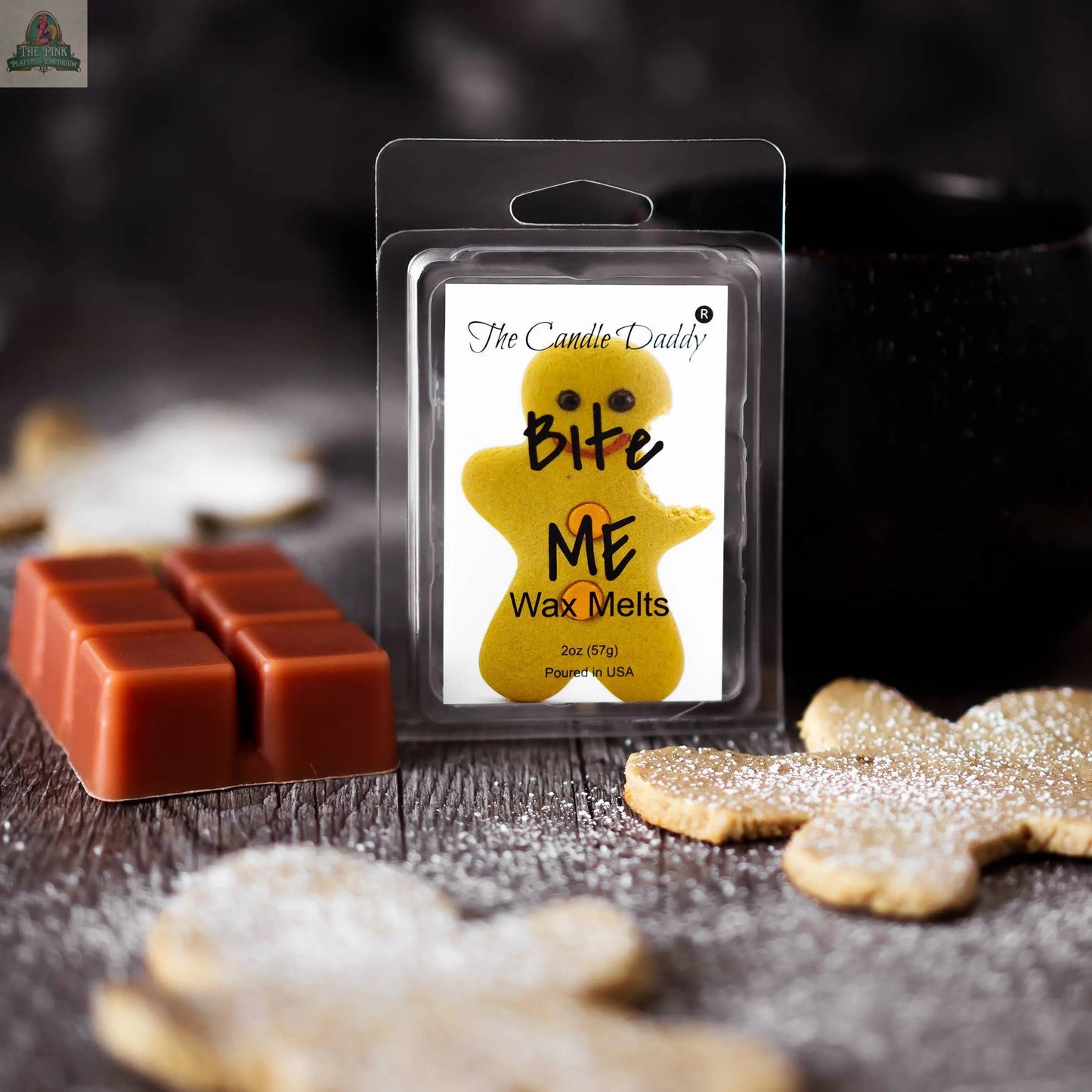 A pack of Bite Me - Funny Christmas Gingerbread Cookie Scented Wax Melts sits on a wooden surface among cookies, a block of wax melts, and a black cup, with powdered sugar scattered around.