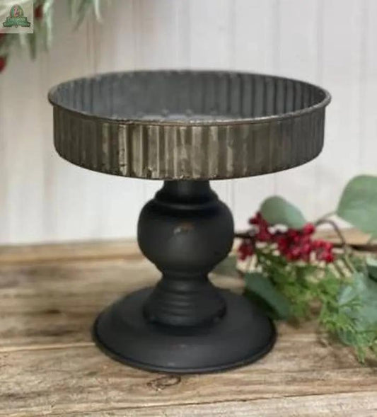The Black Base Candle Pan, featuring a ribbed round tray top and rustic metal design, rests on a wooden surface with greenery and red berries in the background.