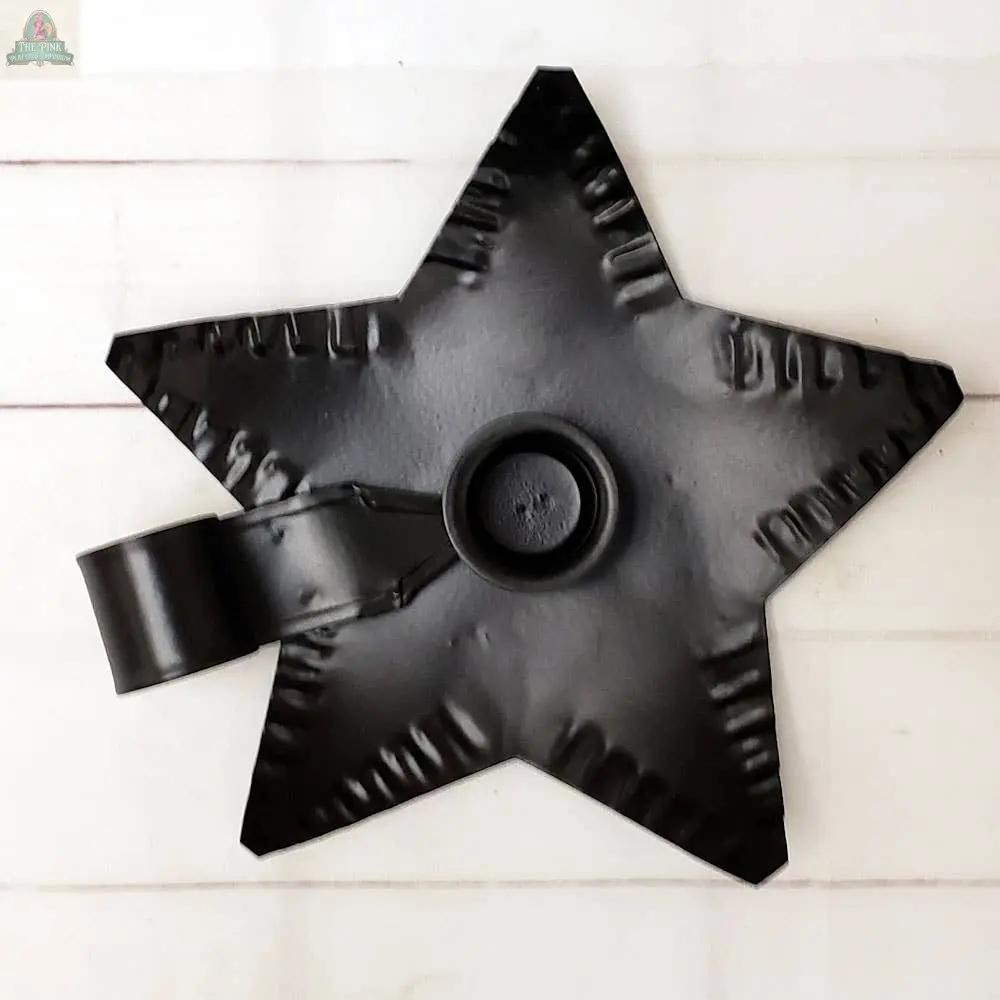 The Black Metal Star Taper Holder features a star-shaped base with a handle and is displayed on a light wooden surface.