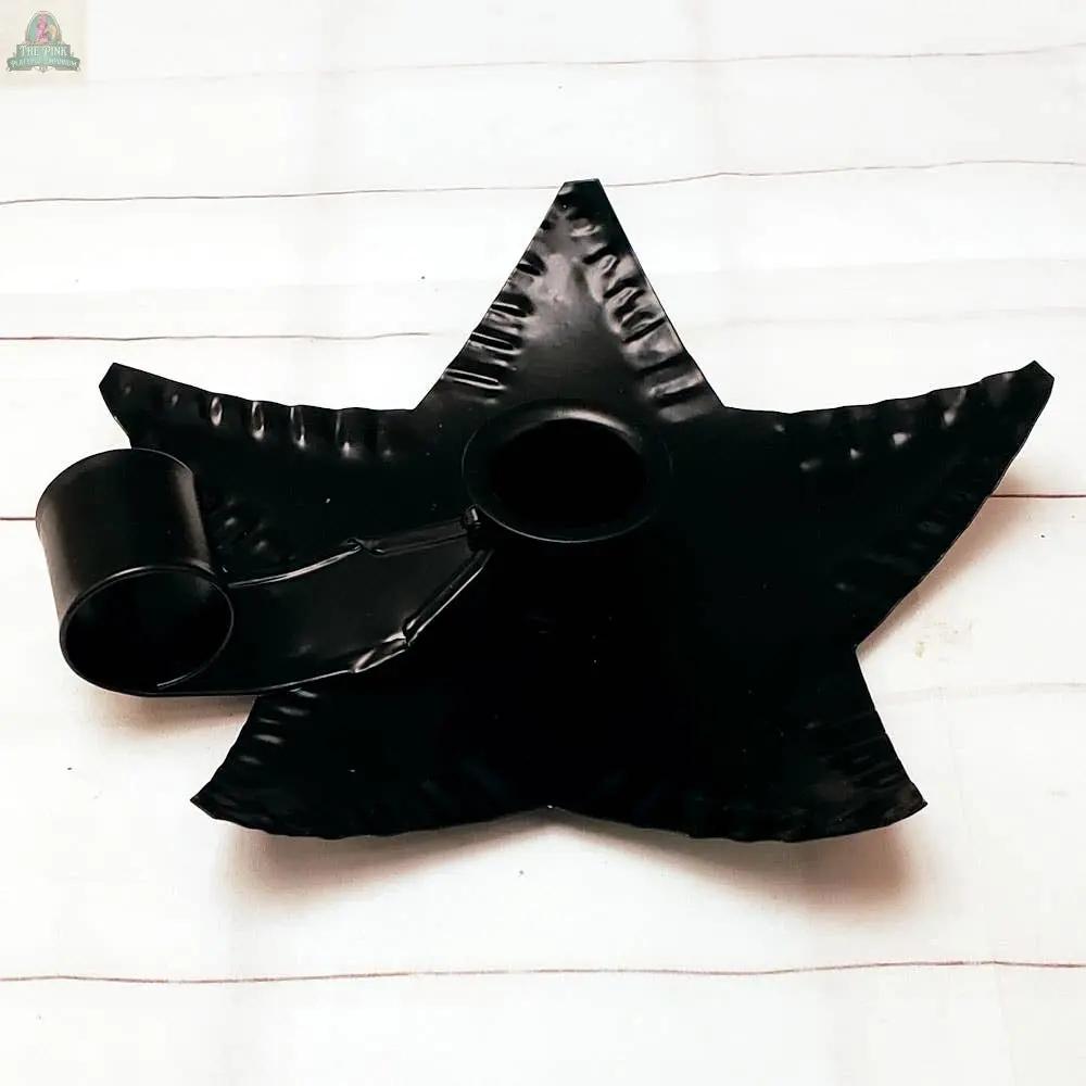 The Black Metal Star Taper Holder, featuring a star-shaped base and round handle, is displayed on a white, lightly striped surface.