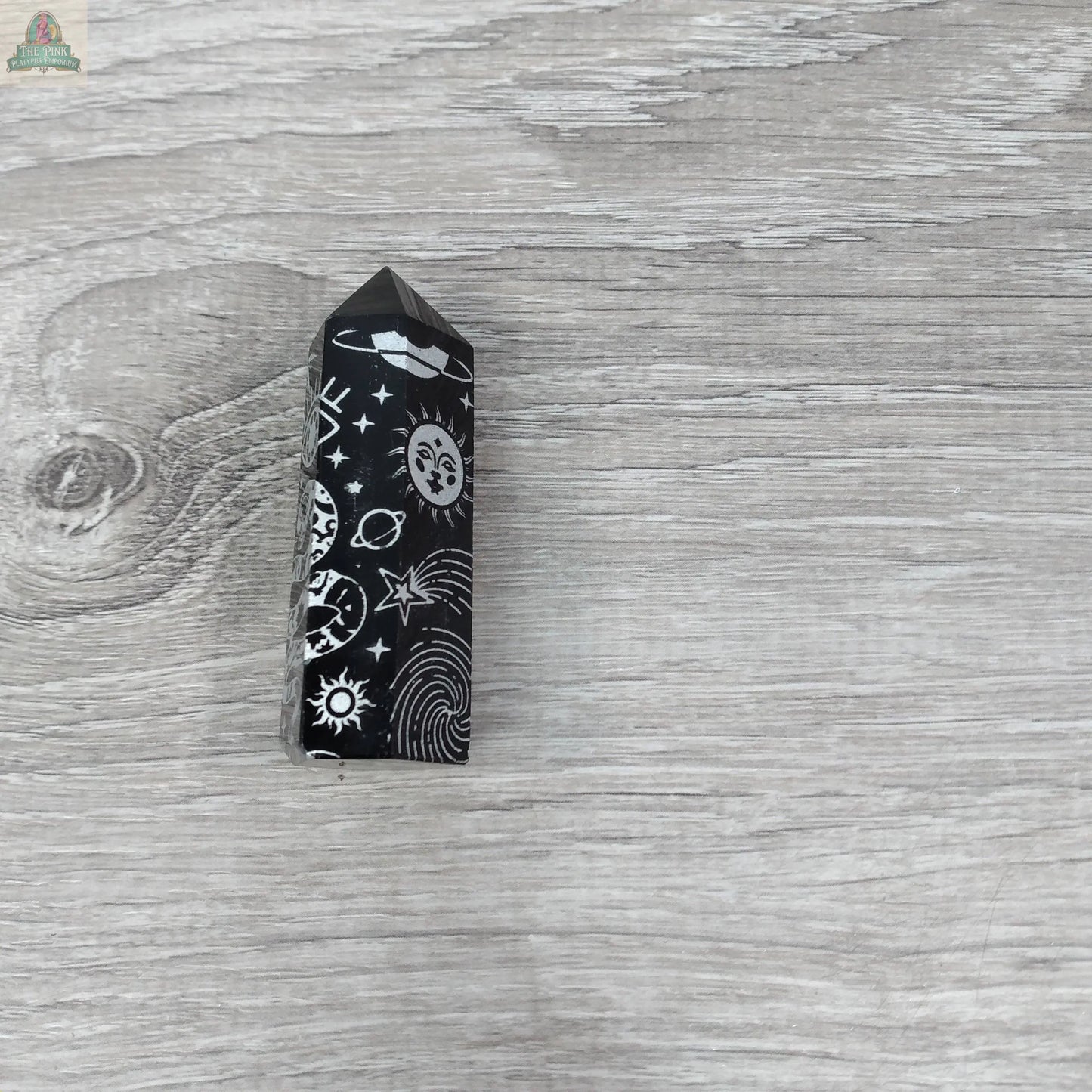 The Keystone Crystals Black Obsidian Chakra Obelisk, etched with celestial designs, rests on a light gray wood surface—an elegant energy work crystal from Pink Platypus Emporium.