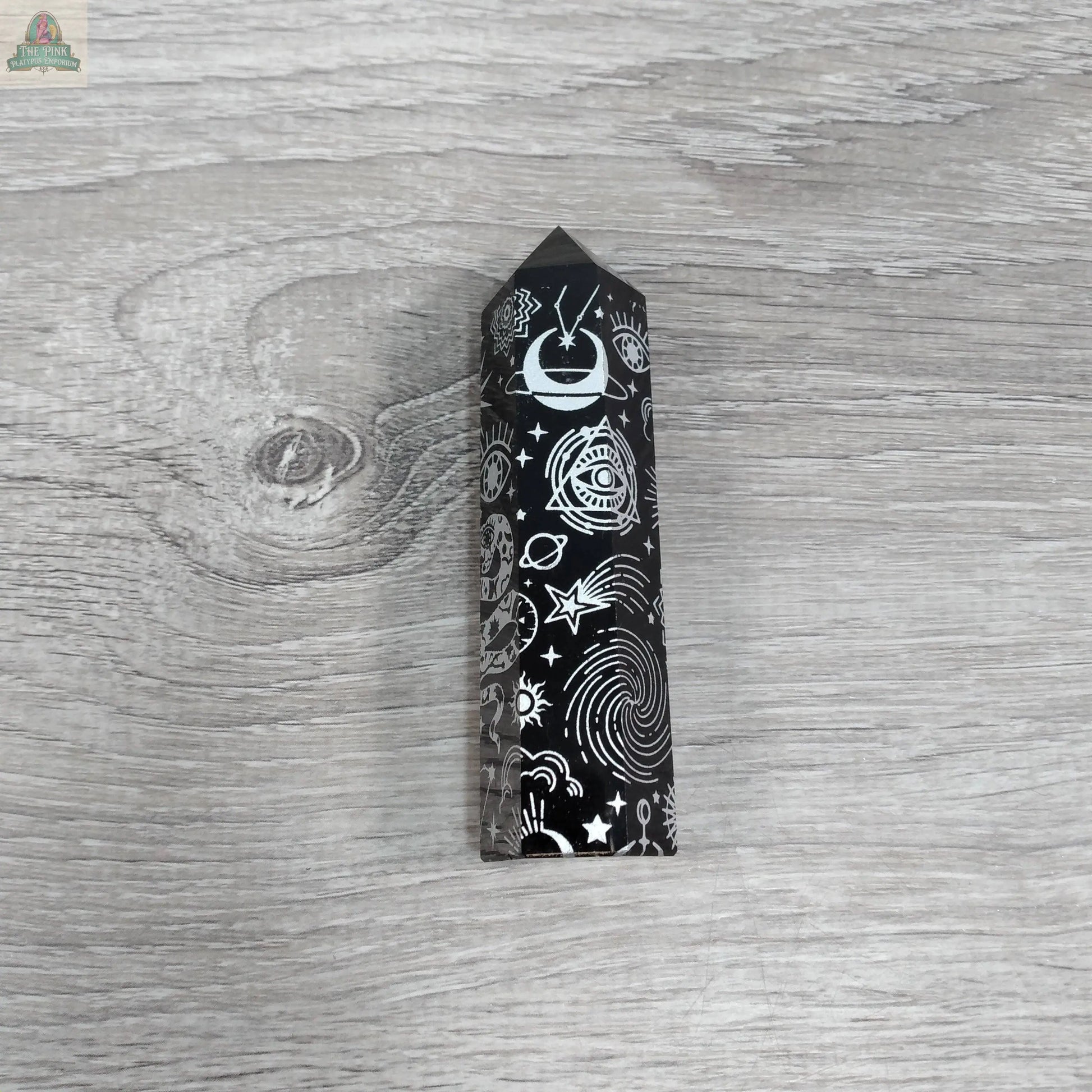 The Black Obsidian Chakra Obelisk by Keystone Crystals, available at Pink Platypus Emporium, features white celestial designs—eyes, stars, moons, and planets—etched on black crystal for energy work and chakra protection.