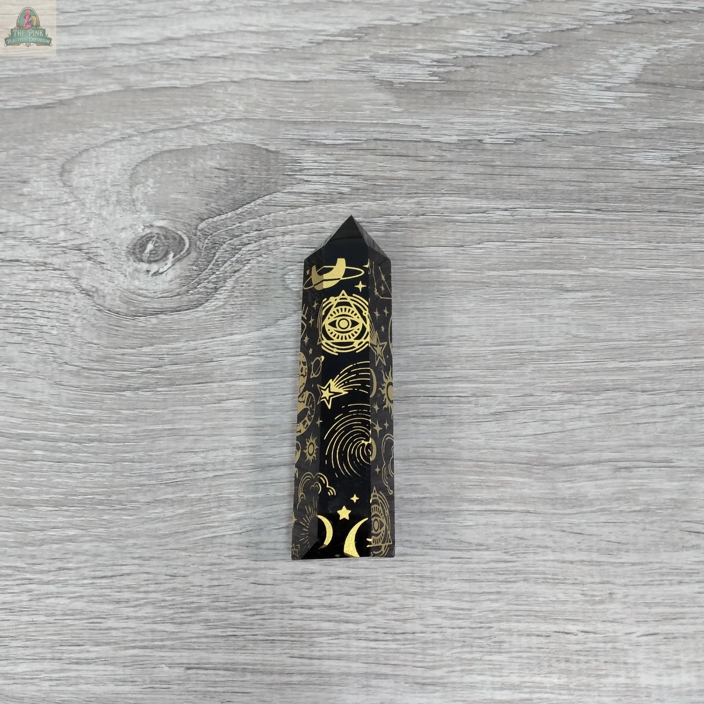 The Keystone Crystals Black Obsidian Chakra Obelisk, etched with gold celestial designs, sits on a gray wooden surface—an energy work crystal from Pink Platypus Emporium.