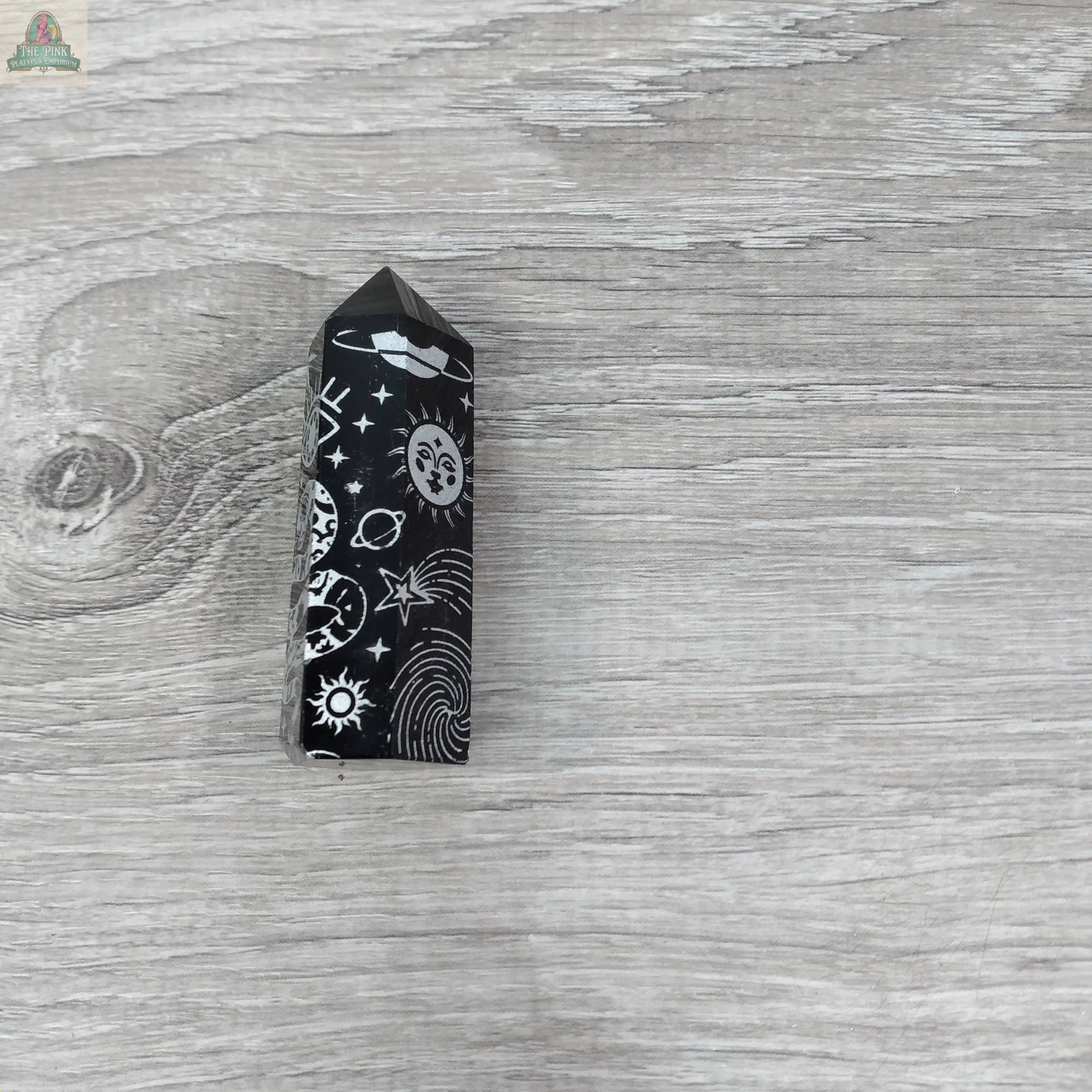 The Black Obsidian Etched Obelisk – Celestial Engraved Crystal Tower by Keystone Crystals features white sun, moon, star, and planet engravings, radiating protective energy and cosmic elegance from Pink Platypus Emporium.