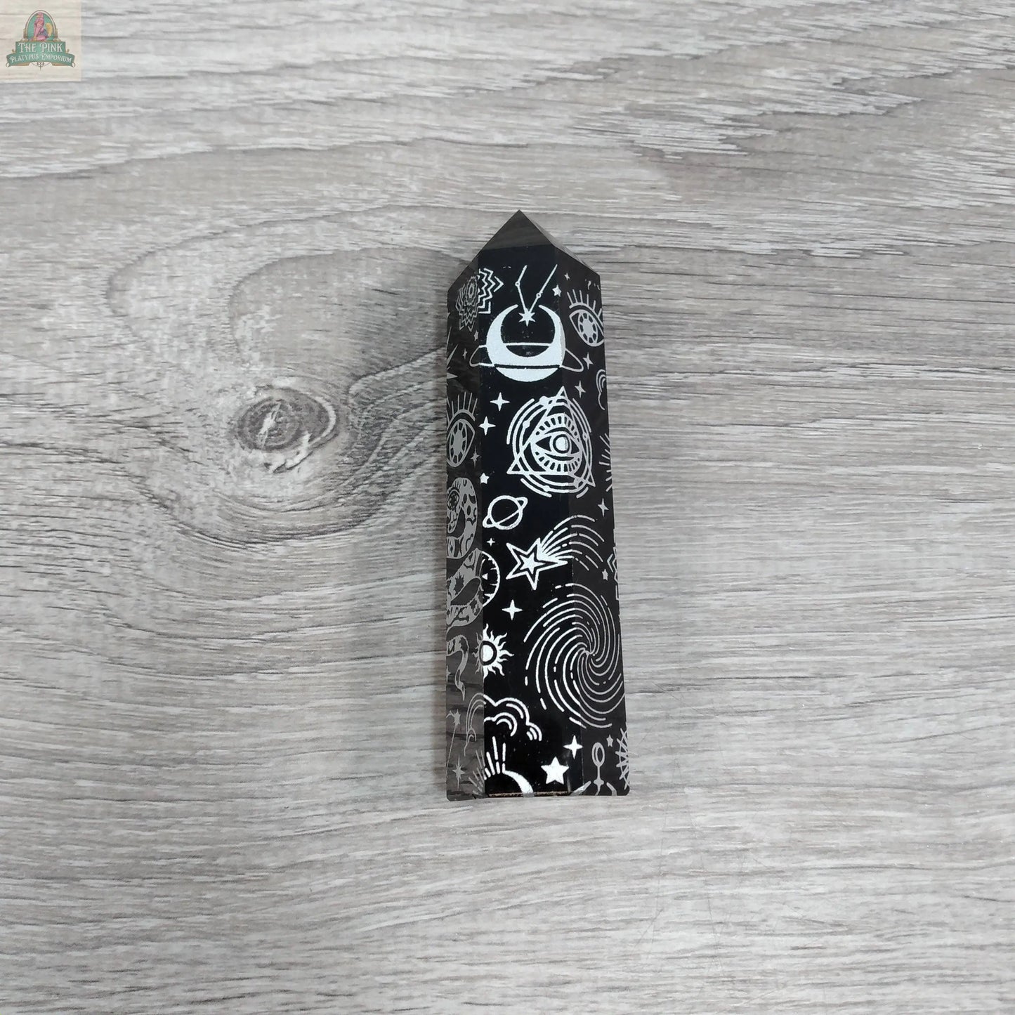 The Keystone Crystals Black Obsidian Etched Obelisk – Celestial Engraved Crystal Tower from Pink Platypus Emporium rests on a light gray wood surface, radiating energy protection with its white celestial engravings.