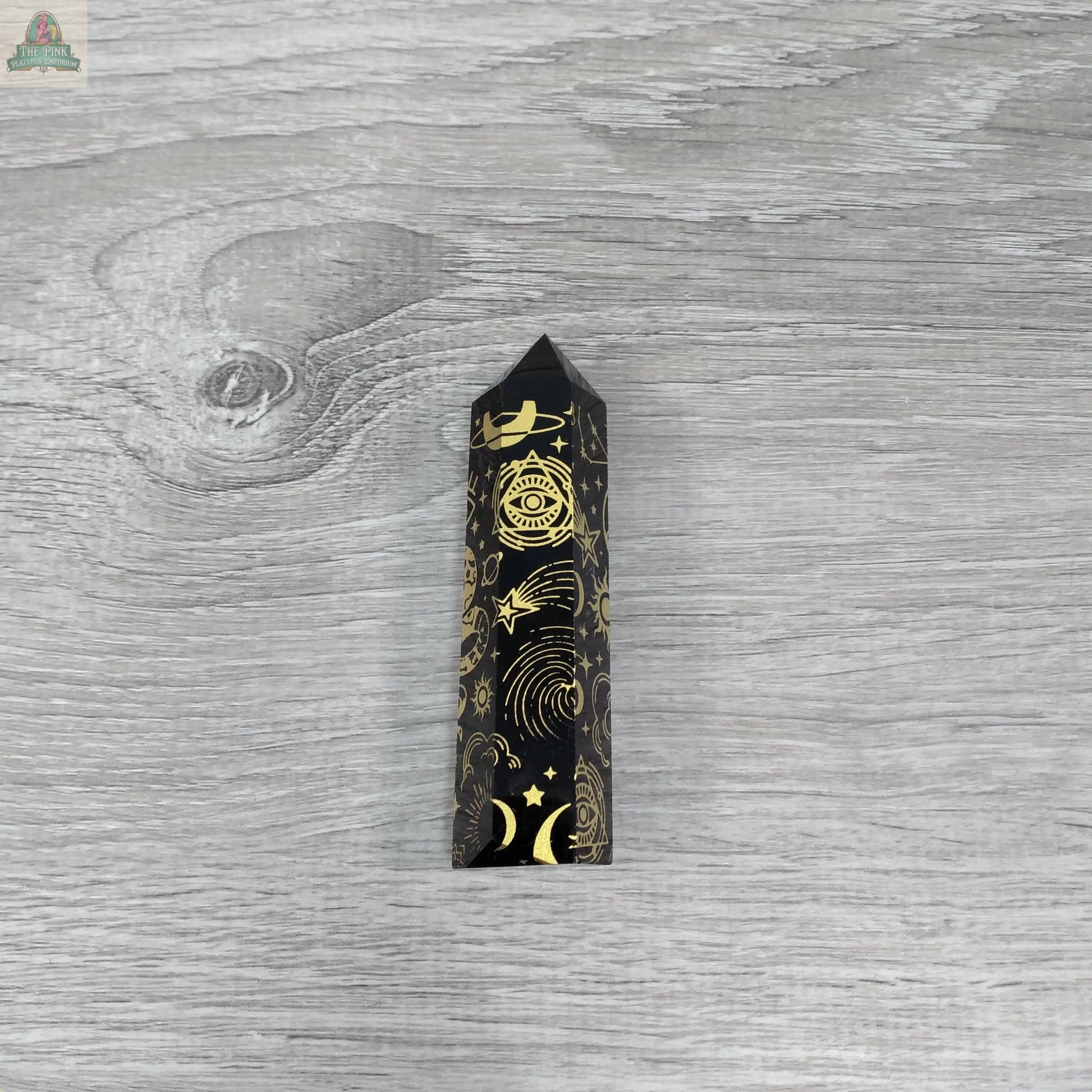 The Black Obsidian Etched Obelisk – Celestial Engraved Crystal Tower by Keystone Crystals from Pink Platypus Emporium rests on a wood surface, radiating energy protection.