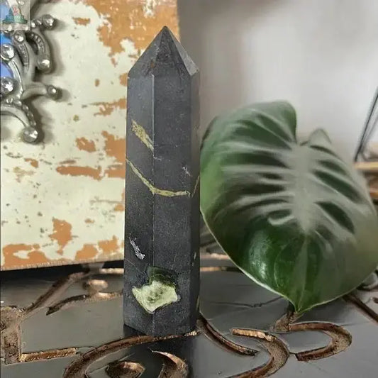 The Black and Green Hematite and Chrysoprase Obelisk Stone Decor features unique greenish streaks and mineral inclusions, displayed beside a large leaf and a textured background—ideal for crystal-themed home decor enthusiasts.