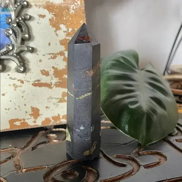 The Black and Green Hematite and Chrysoprase Obelisk Stone Decor stands upright on a carved surface beside a large green leaf and a distressed ornate mirror, creating an elegant, polished crystal accent for any space.