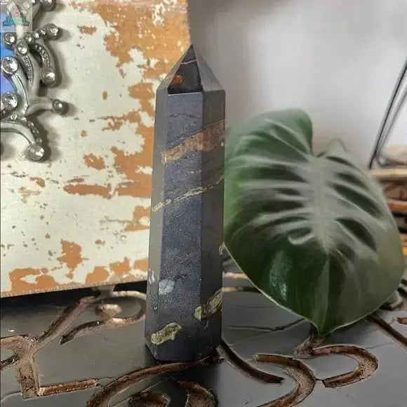 A Black and Green Hematite and Chrysoprase Obelisk Stone Decor stands upright on an ornate surface, with a large green leaf and a distressed tan and white decorative panel displayed behind it.