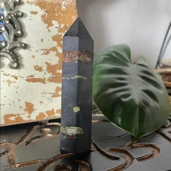 The Black and Green Hematite and Chrysoprase Obelisk Stone Decor stands upright on a surface, adding striking crystal decor, with a large green leaf and a distressed painted object in the background.